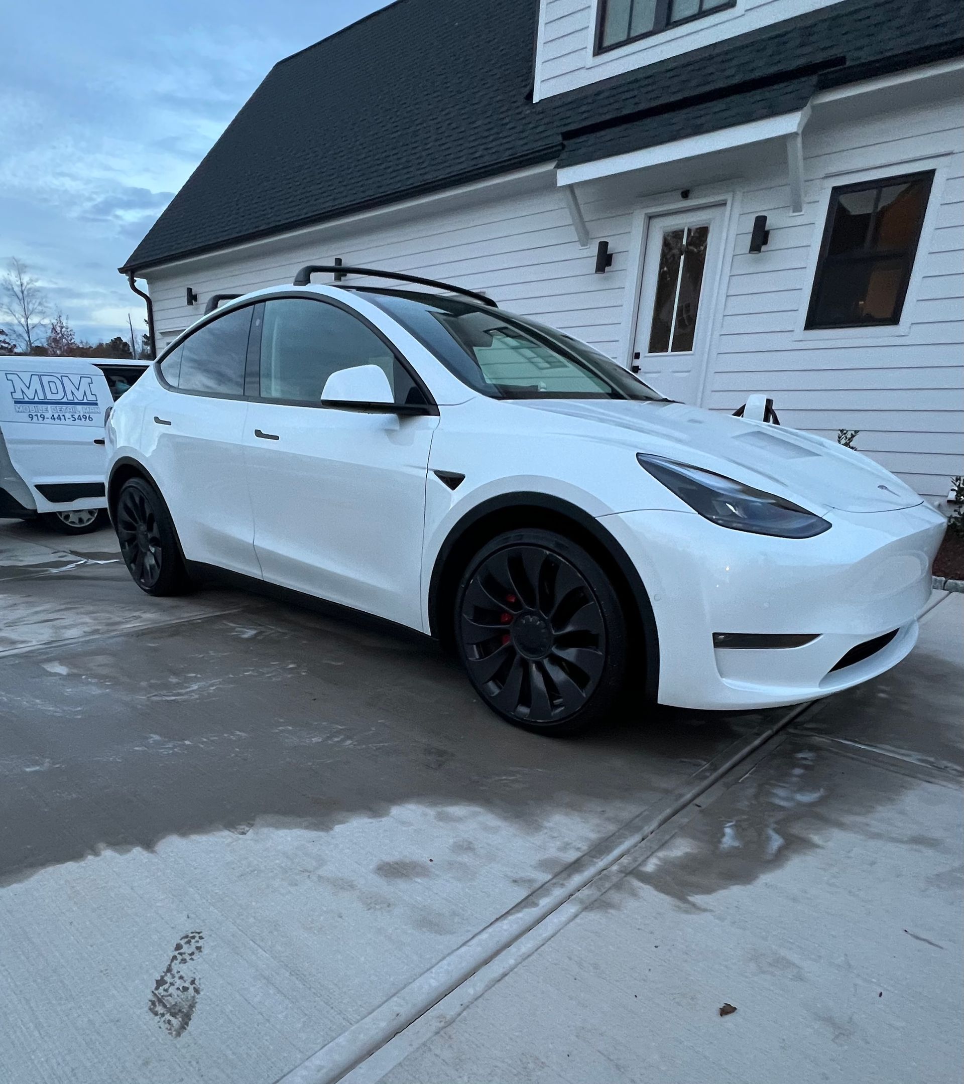 White Tesla Y, parked on wet pavement in front of a white house with black trim.