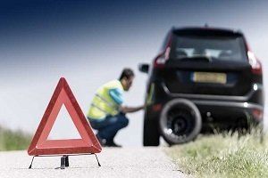 Car with flat tire on roadside; person changing tire, warning triangle in foreground.