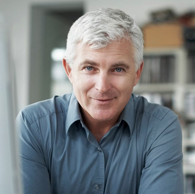 Man with gray hair, blue shirt, smiling at camera. Blurred office setting.