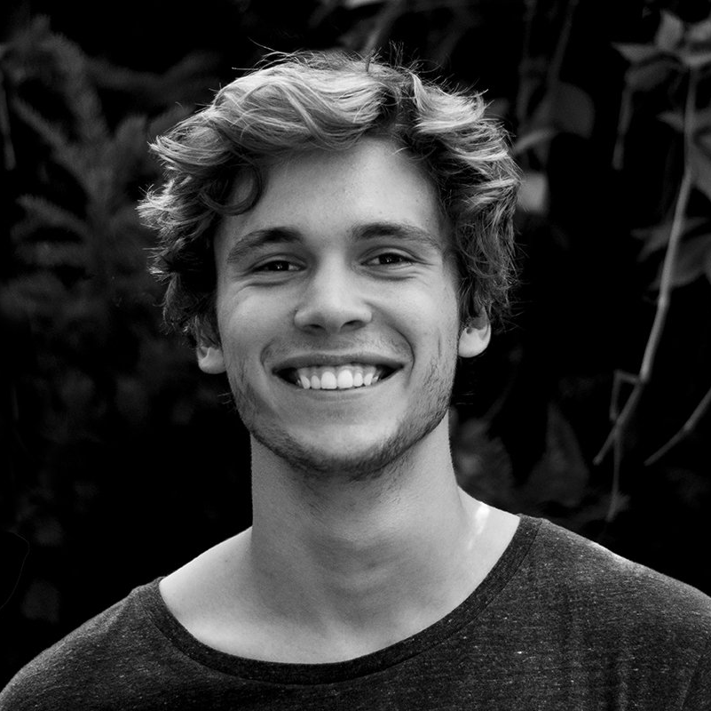 Smiling man with curly hair, wearing a dark shirt, in a natural setting.