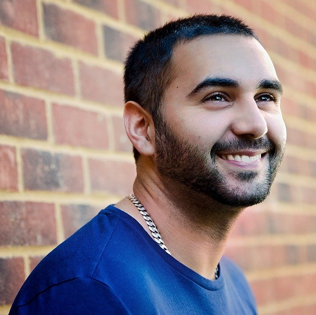 Man with dark hair and a beard smiles, looking upward near a brick wall, wearing a blue shirt.