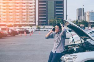 Man on phone with car hood up, appears distressed in a parking lot.