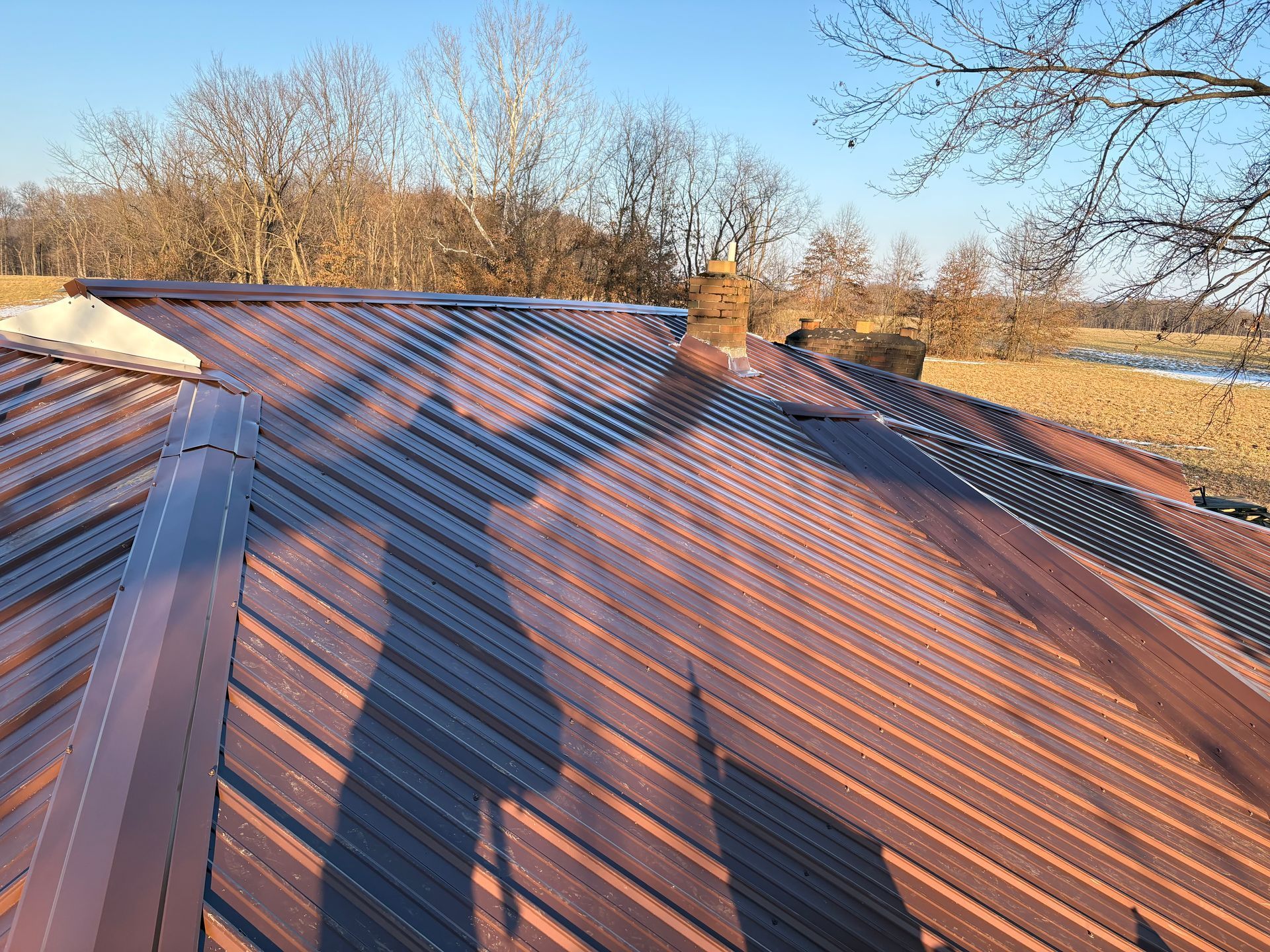Brick house with metal roof under a gray sky, surrounded by leafless trees.