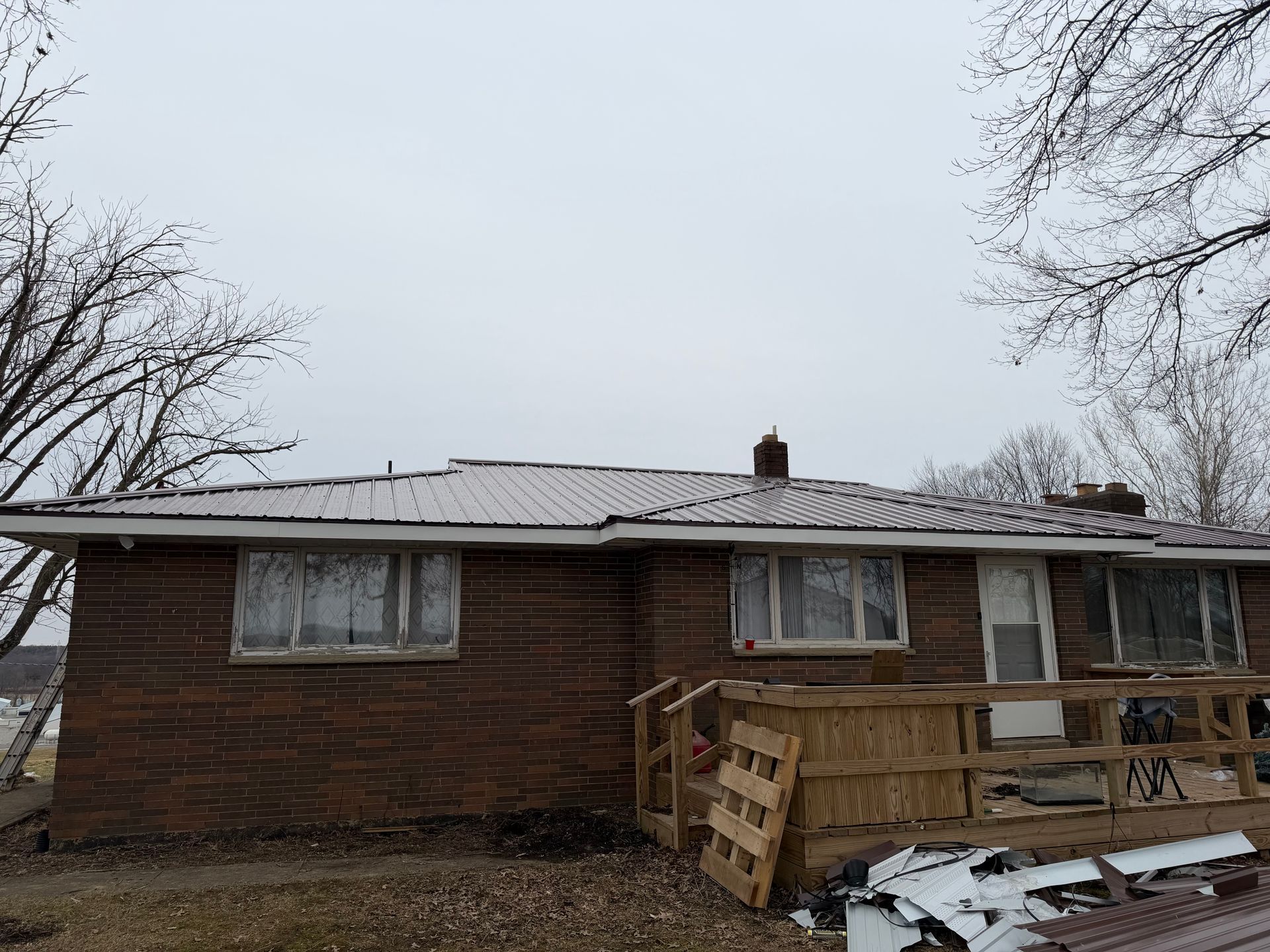 Brick house with metal roof under a gray sky, surrounded by leafless trees.