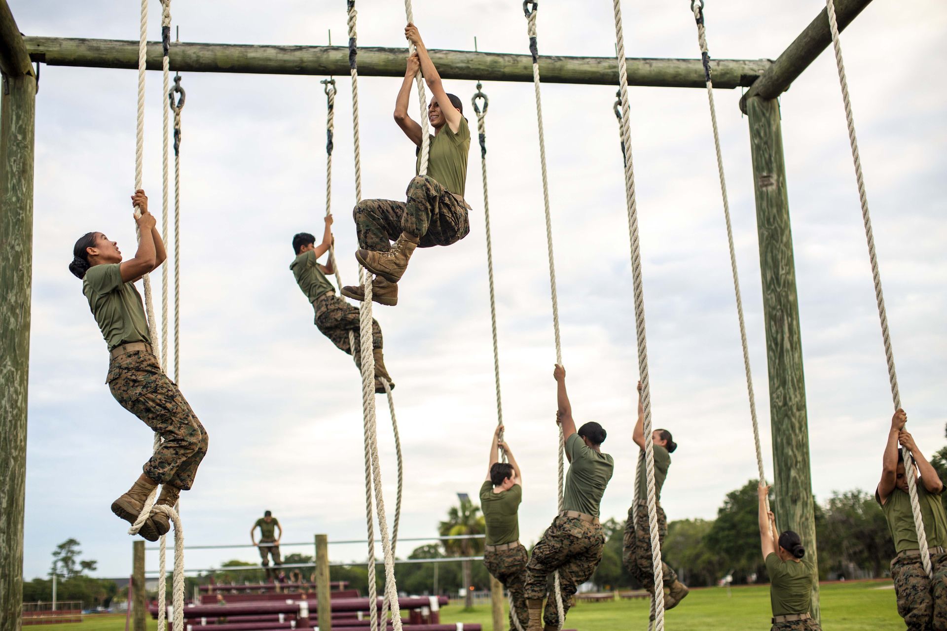 Marines in green uniforms climbing ropes on an outdoor obstacle course.