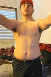 Man with arms outstretched, shirtless, wearing a red cap and black shorts. Indoor setting.