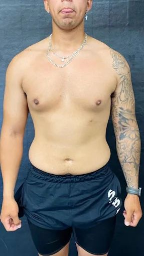 Man with tattoos on right arm, wearing black shorts and necklace, posing shirtless, in a gym.