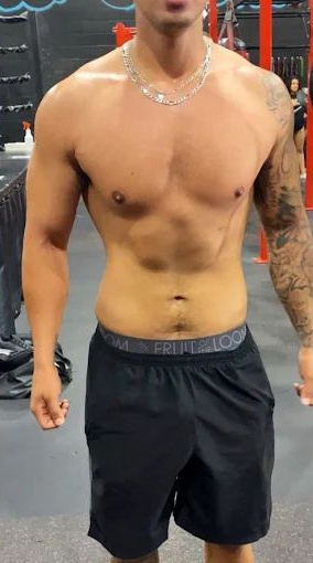 Man in gym, shirtless with arm tattoo, wearing black shorts, silver chain.
