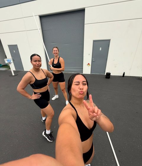 Three women in black workout clothes pose outside, one taking a selfie, others smiling and winking.