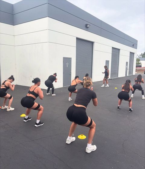Group of women doing squats outdoors, wearing workout clothes with resistance bands.