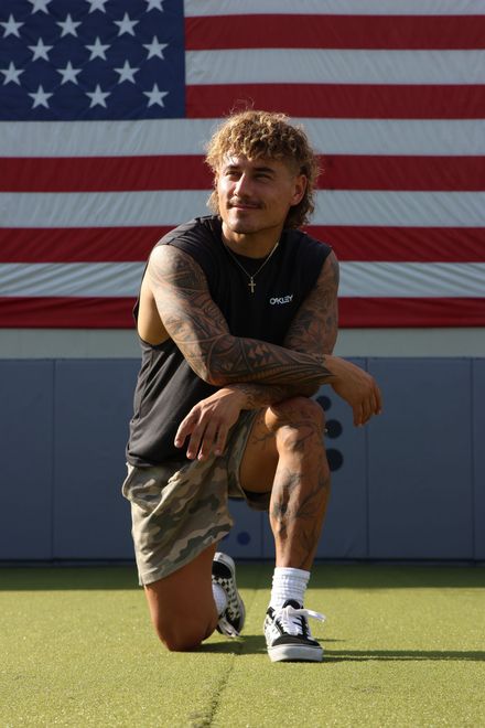 Man kneeling on a green surface with American flag backdrop, wearing sleeveless shirt, shorts, and sneakers.
