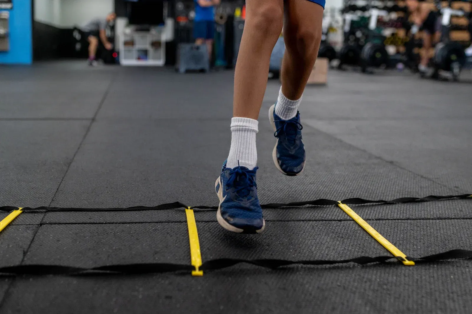 Person in blue shoes and white socks running agility ladder in a gym.