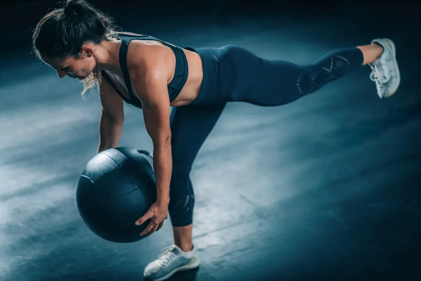 Woman balances on one leg, bending over a medicine ball in a dark studio, workout.
