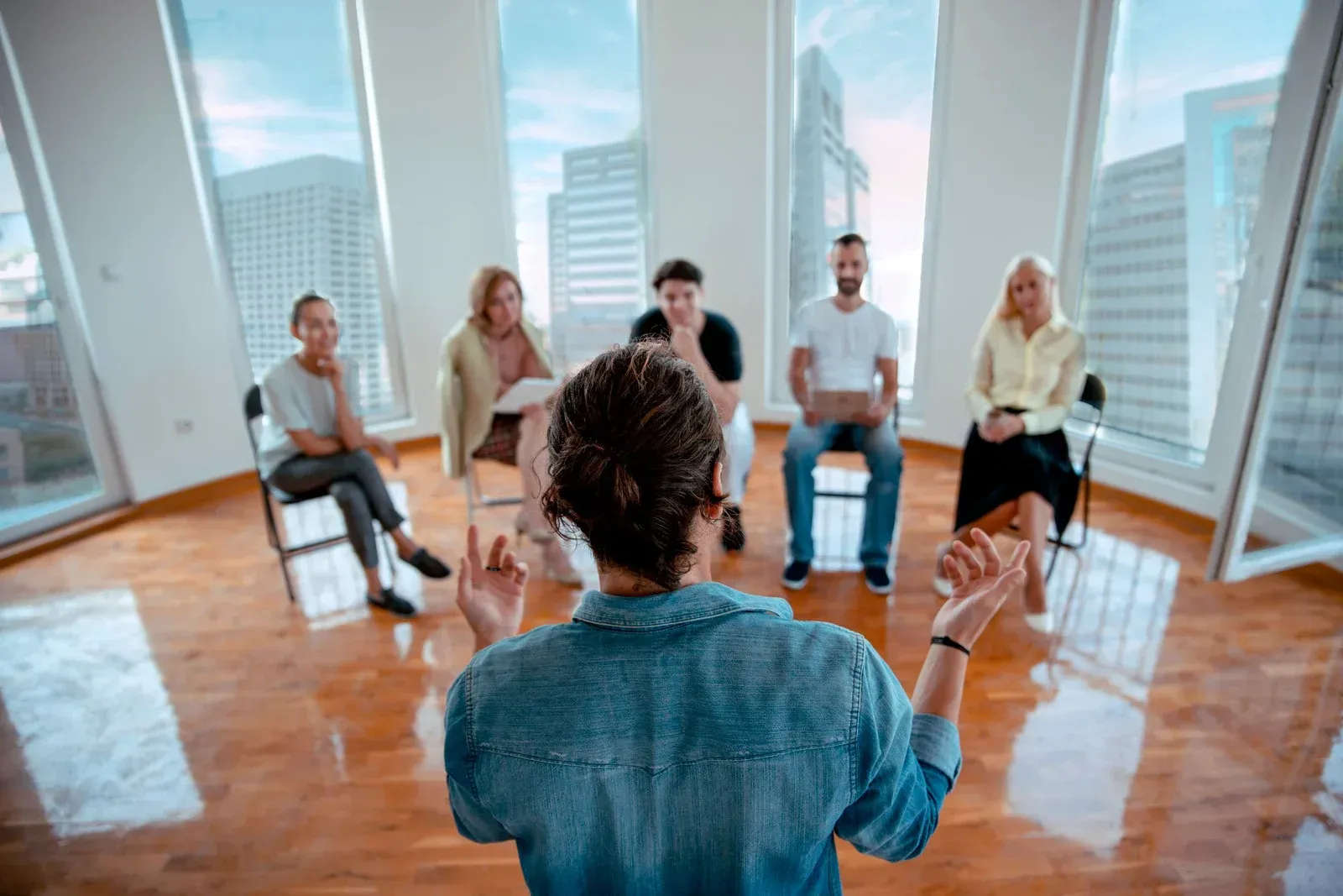 A person gestures to a seated group in a bright room with city views.
