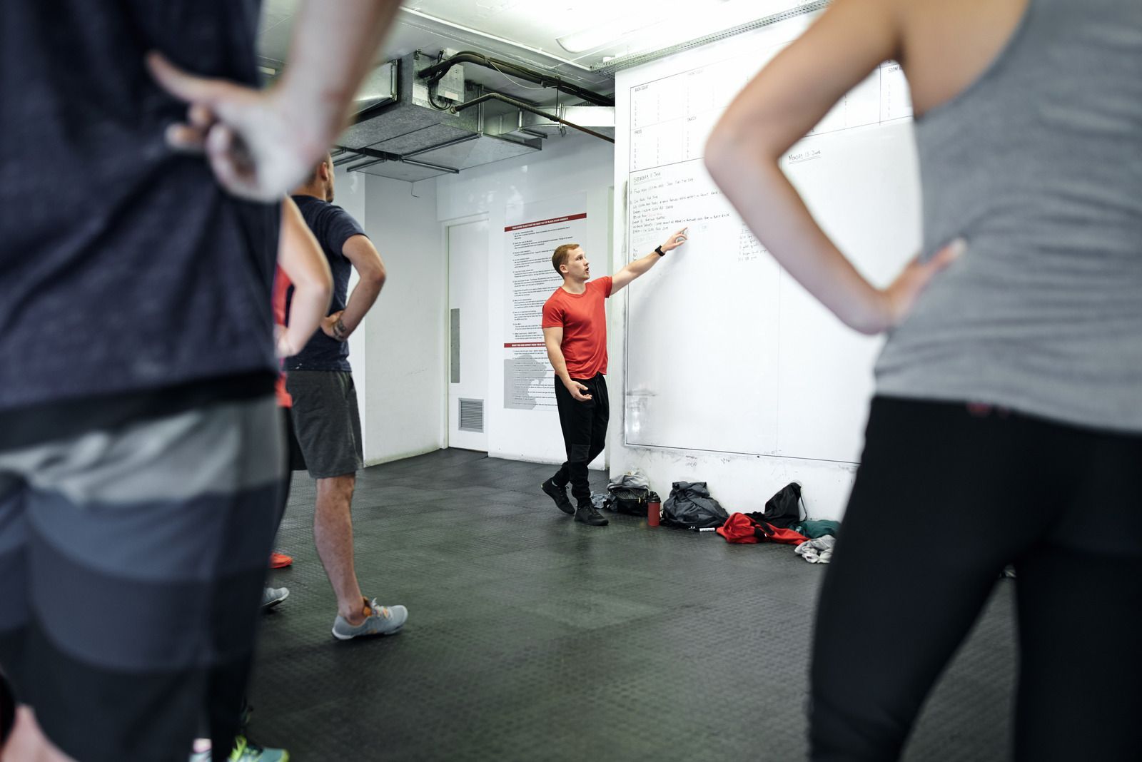Group of people in workout clothes watch a fitness instructor point to a whiteboard in a gym.