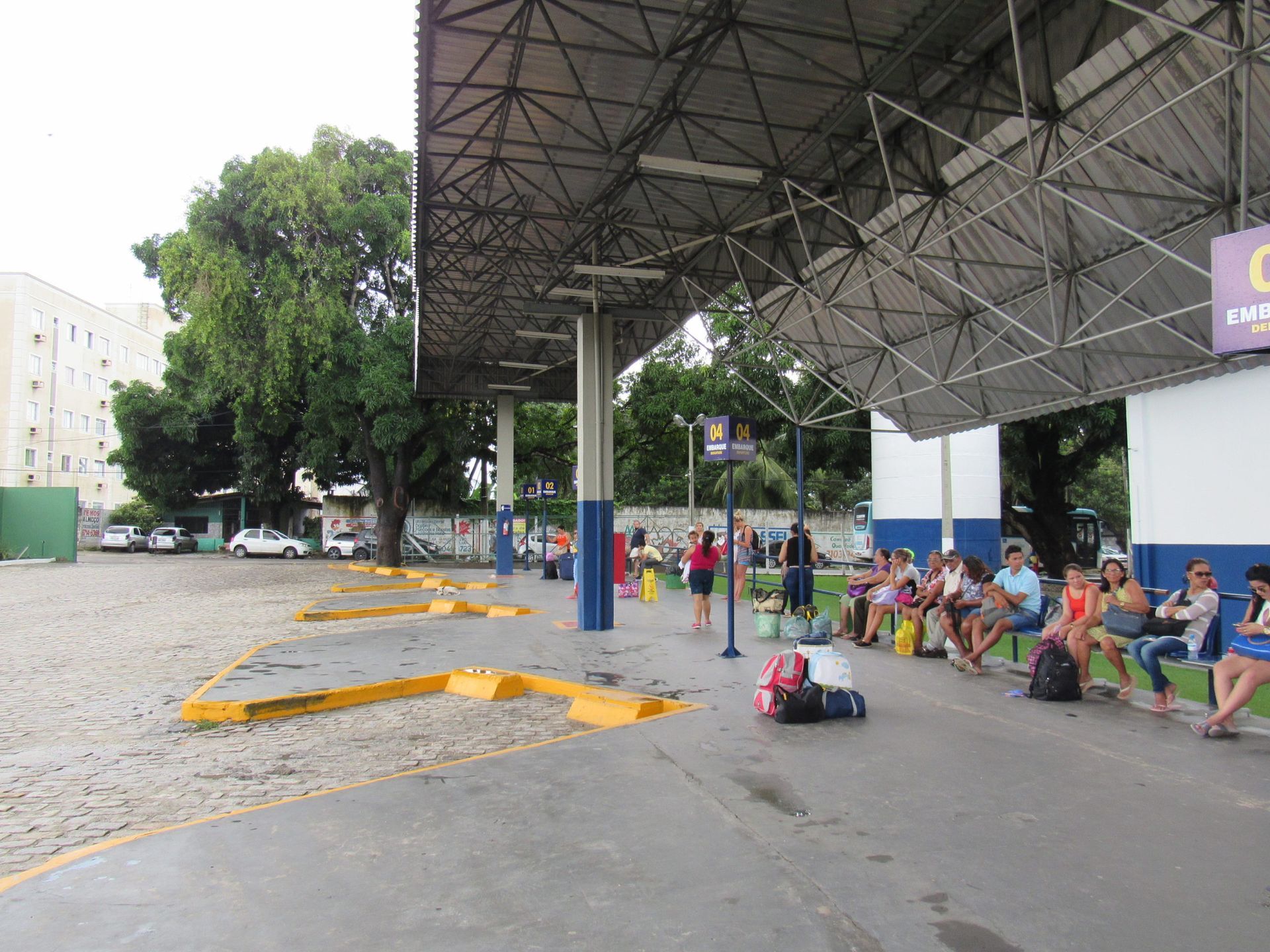 Estação rodoviária, pessoas esperando sob um telhado de metal, com bagagens.