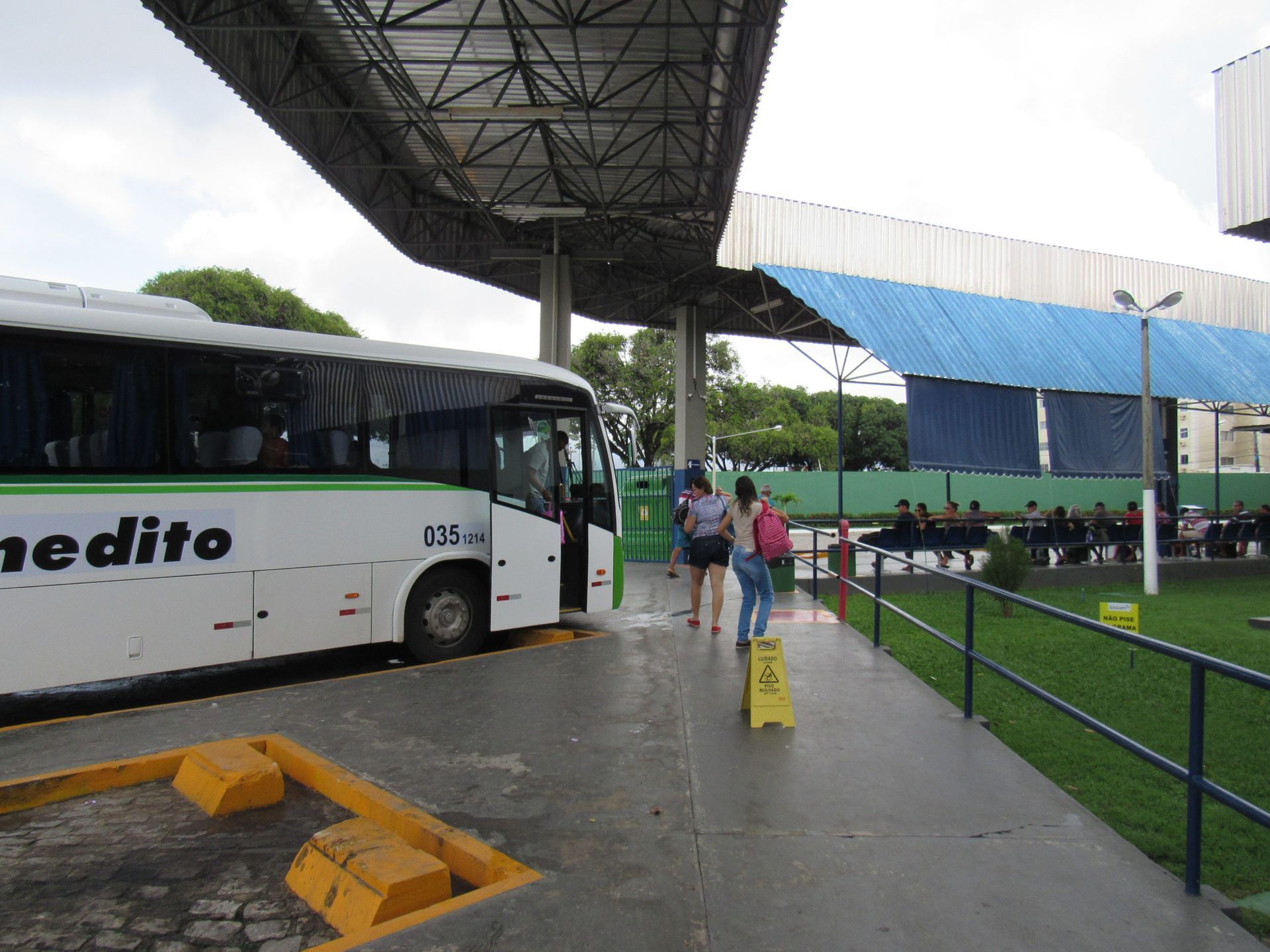Um ônibus estacionado sob um abrigo. Passageiros embarcam/desembarcam. Passarela de concreto, gramado verde, telhado azul.