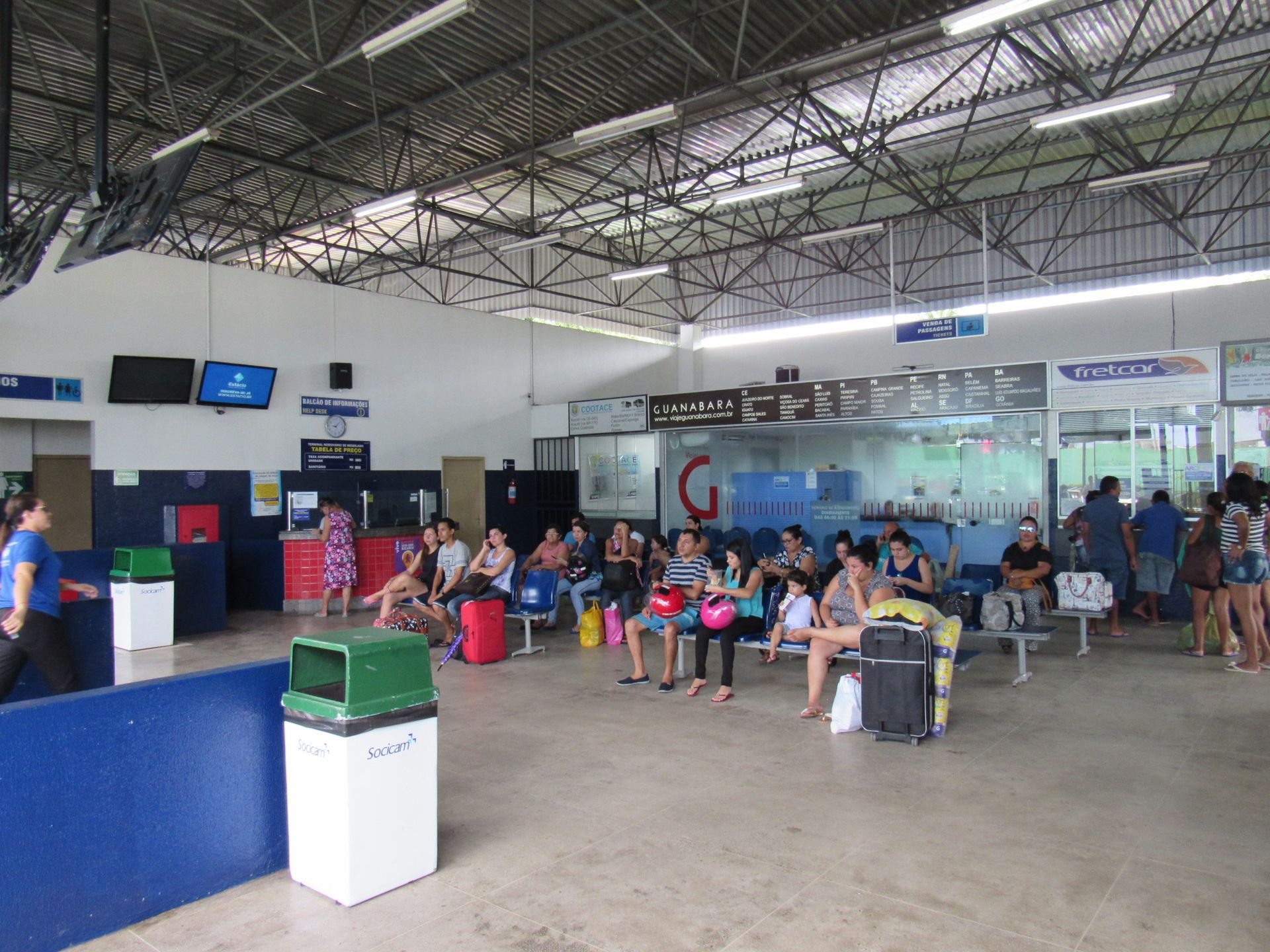 Pessoas aguardando em um terminal de aeroporto bem iluminado. Bancos, lixeiras e informações sobre voos são visíveis.