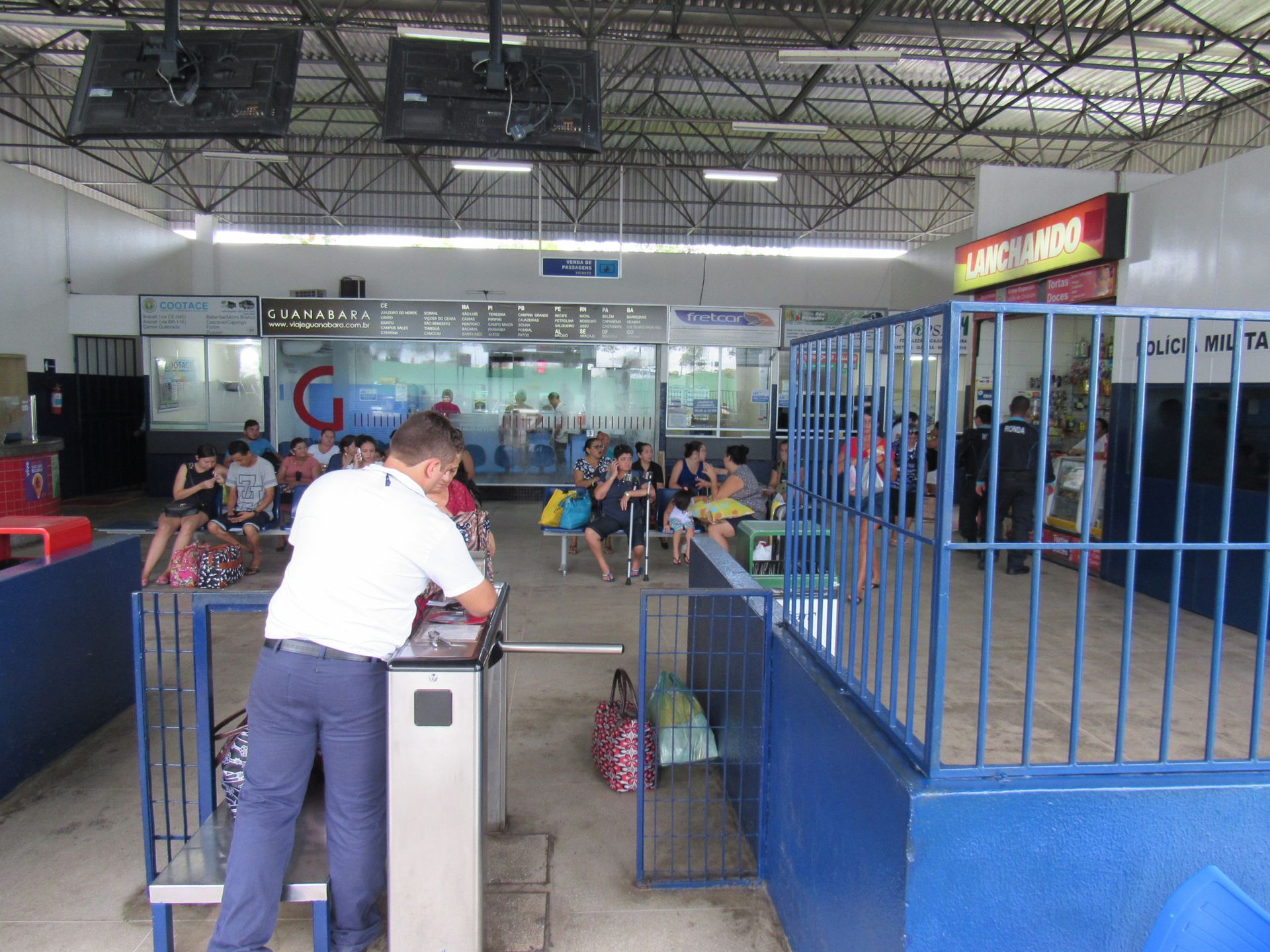 Um homem uniformizado confere os bilhetes em uma catraca na área de espera. Ao fundo, pessoas estão sentadas às mesas.