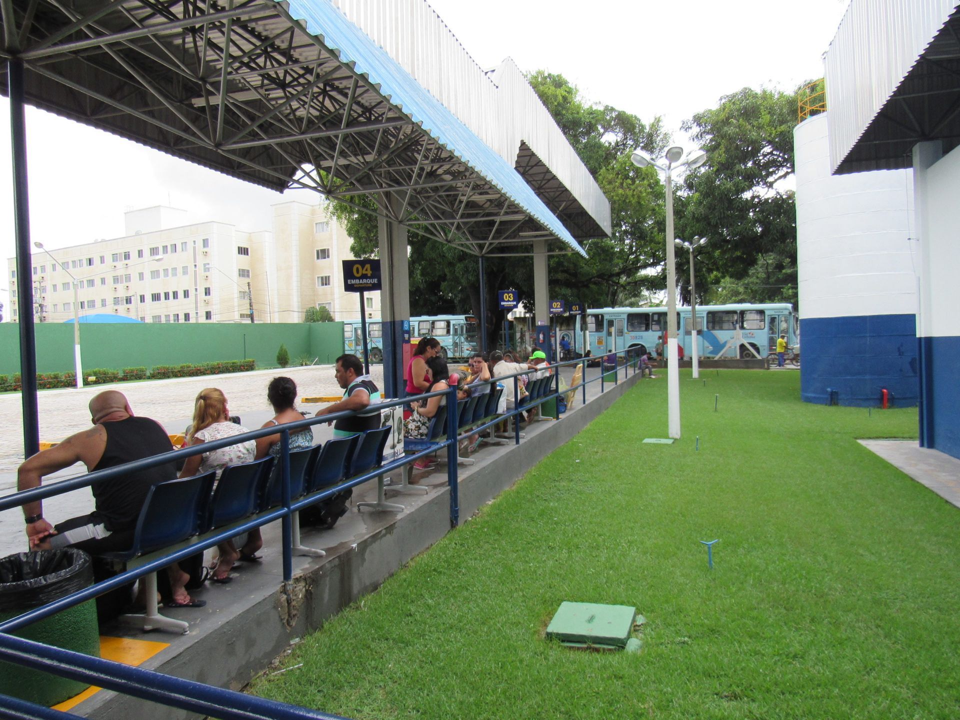Pessoas aguardam em um ponto de ônibus com assentos azuis, sob uma área coberta. Grama, ônibus e um prédio são visíveis.
