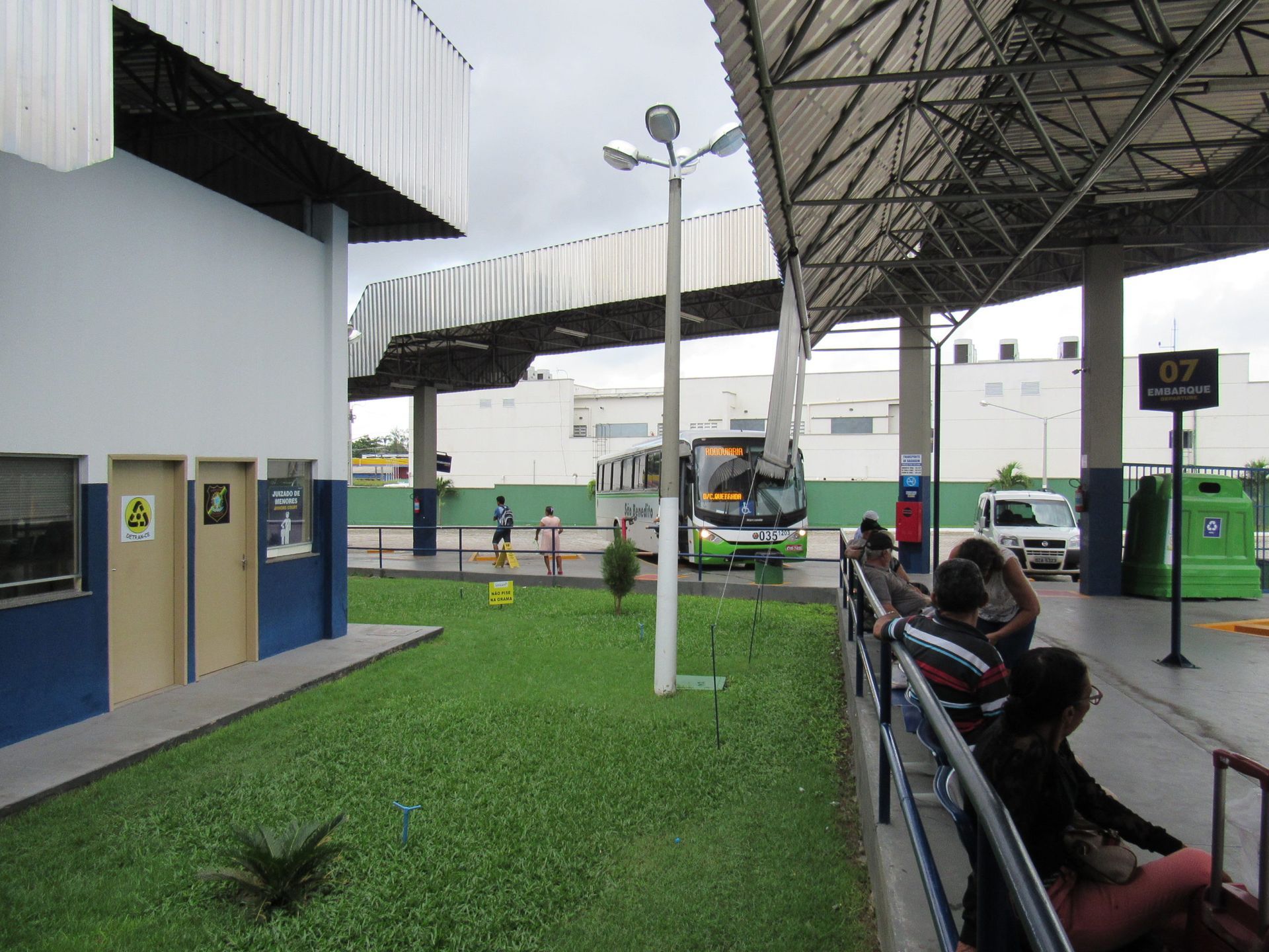 Placa do terminal rodoviário de João Pessoa, com uma fila de carros brancos, sob um céu nublado.