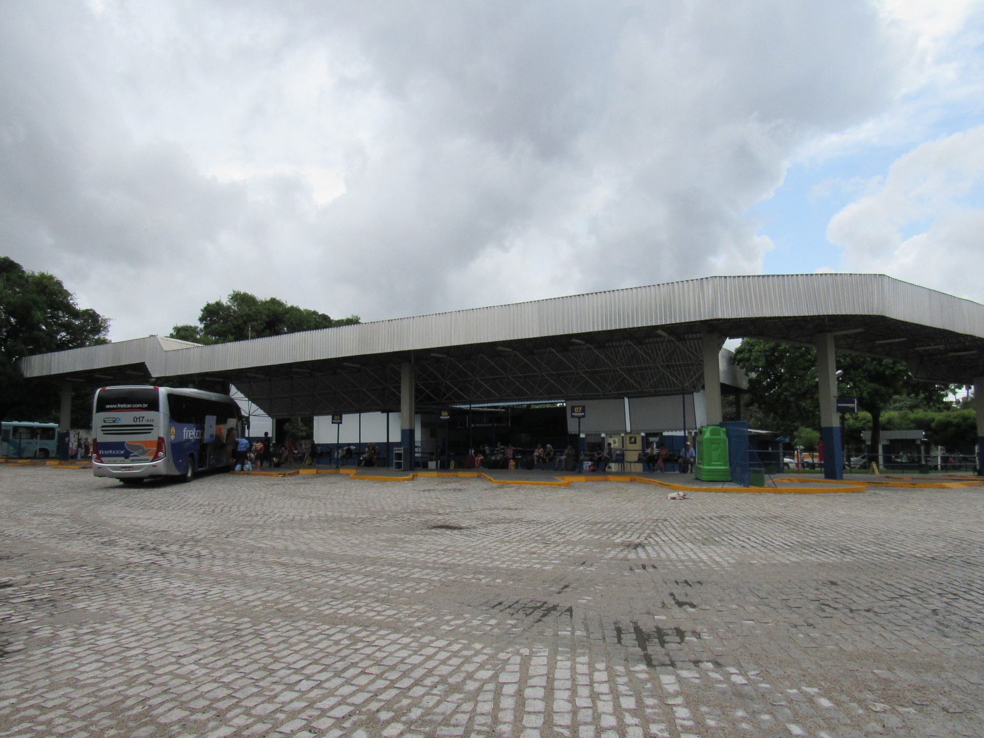 Táxis brancos enfileirados sob a cobertura de um prédio; plantas verdes e um céu azul se destacam.