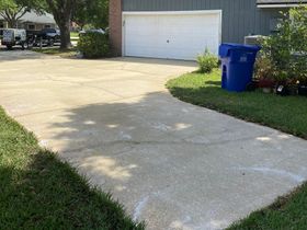 A concrete driveway leading to a garage with a blue trash can on the side of it.