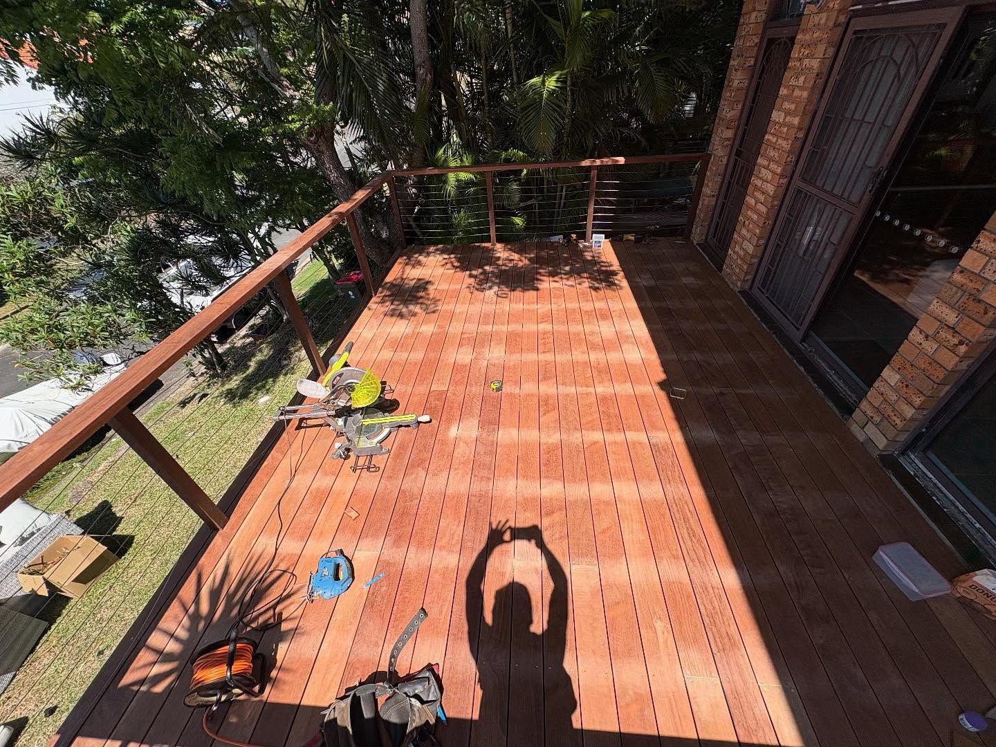 Wooden deck with railing and trees, sunlit with scattered tools and a person’s shadow taking a photo