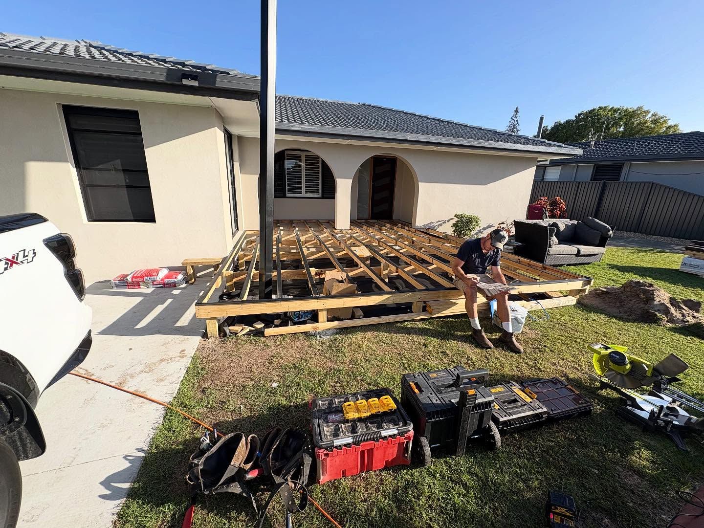 Workers building a wooden deck in a front yard beside a white house, with tools and lumber laid out.