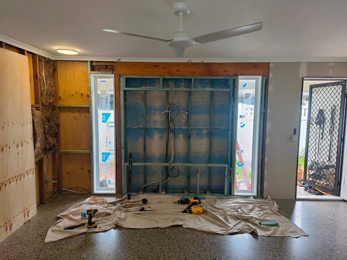 Living room under renovation with covered floor, exposed walls, and blue-framed window at center