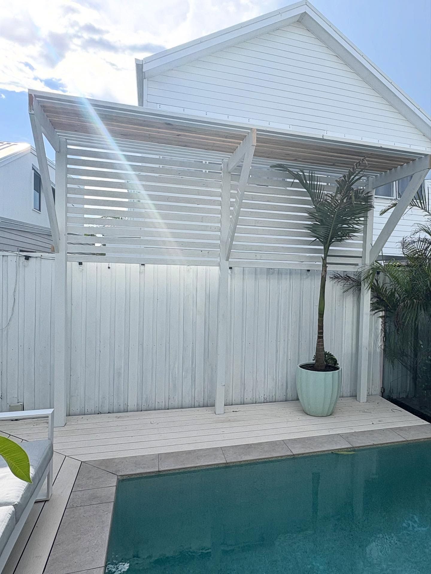 White pergola beside a turquoise pool, with a white fence and potted tree in a backyard patio area