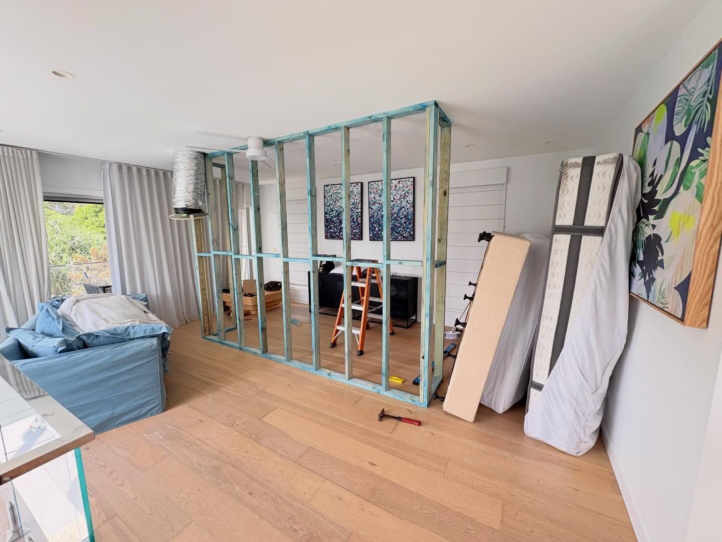 Partially renovated living room with exposed wall framing, blue sofa, and artwork on white walls