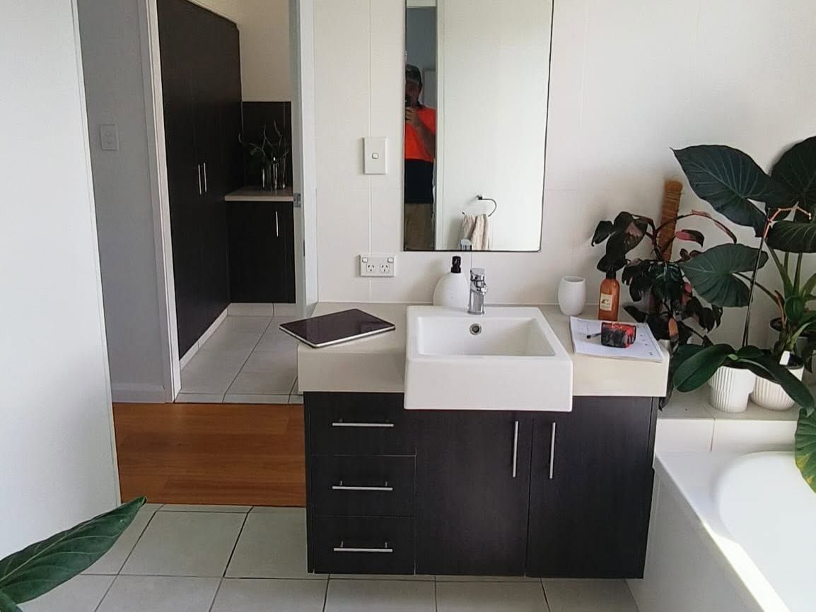 Modern bathroom vanity with dark cabinets, white sink, mirror, and large plant by the tub.
