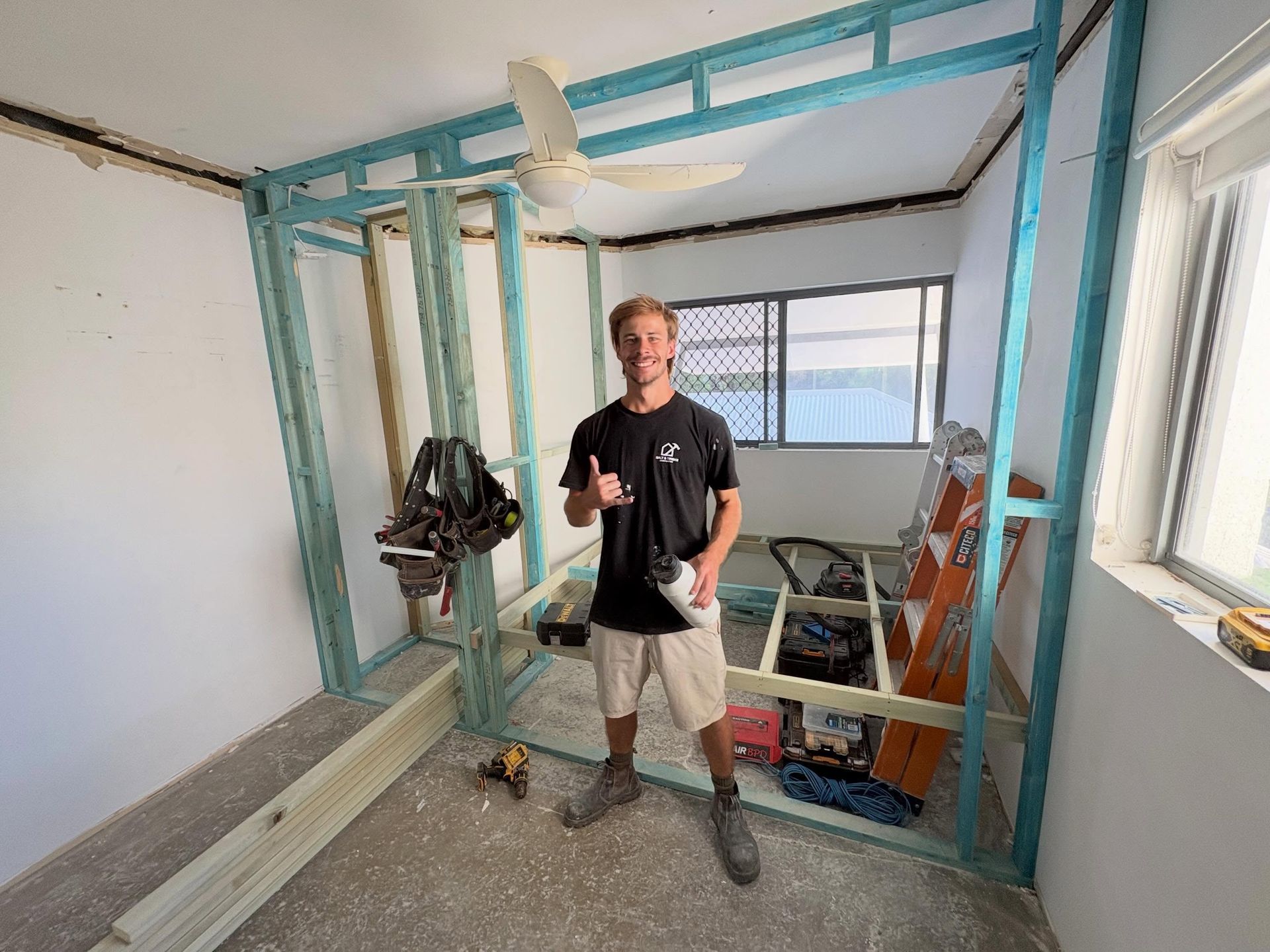 Man standing in an unfinished room beside a blue-framed wall structure with tools and construction materials.