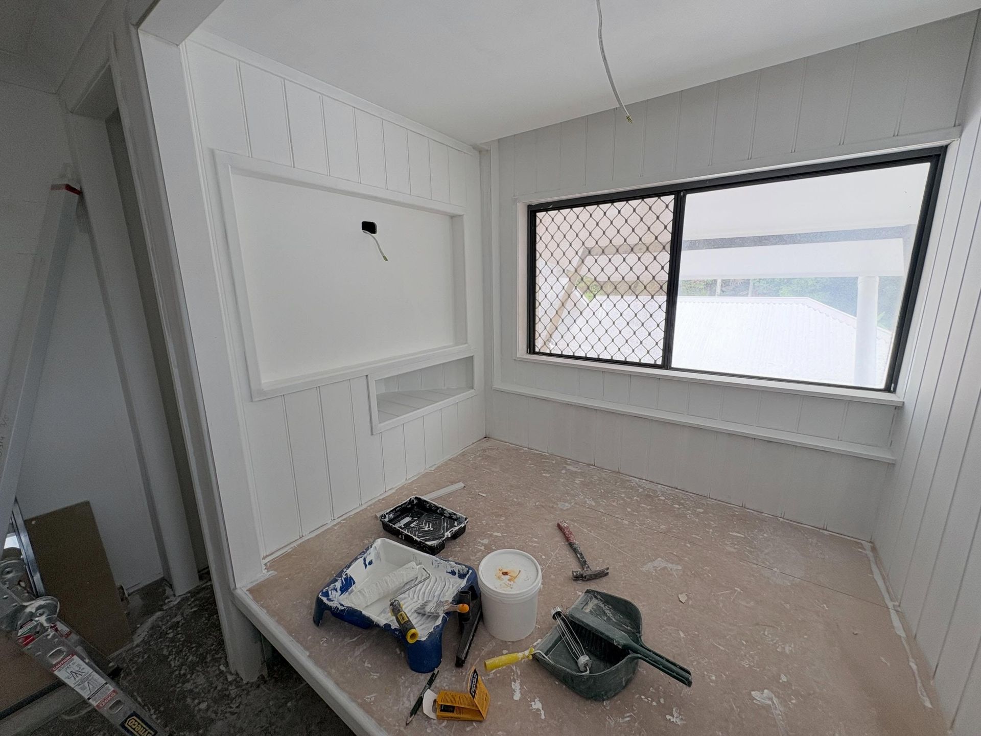 Small room under renovation with tools and paint supplies on the floor, white walls, and a window.