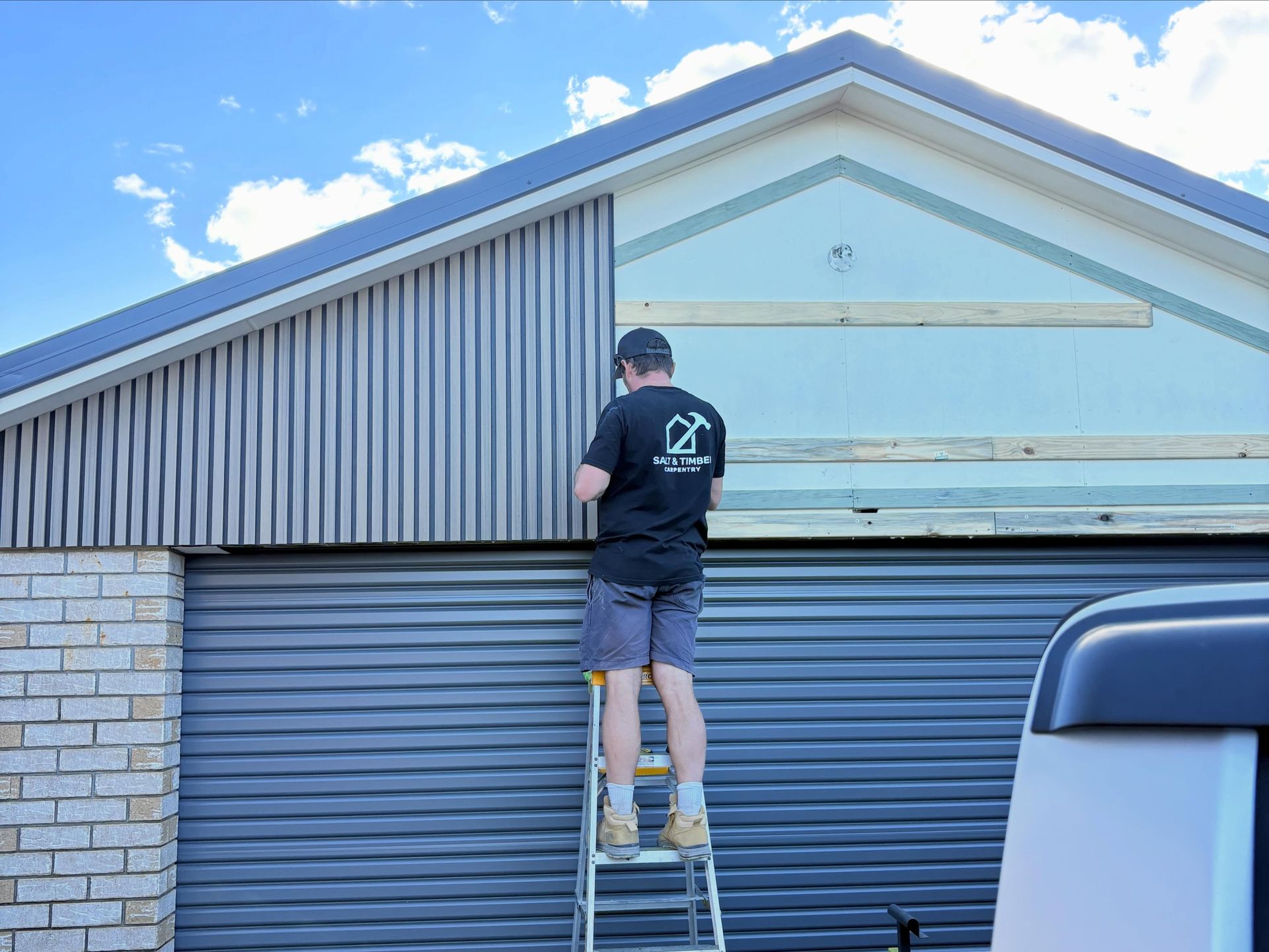 Person on a ladder painting the front gable of a house above a blue garage door.