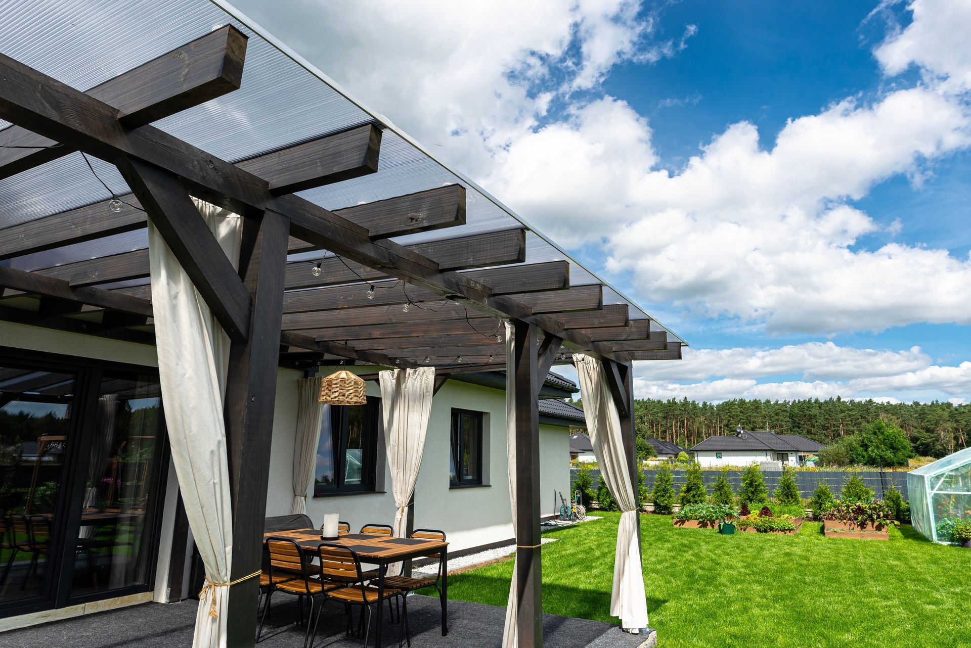 Modern patio with pergola, outdoor dining table, white curtains, and green lawn under a cloudy blue sky