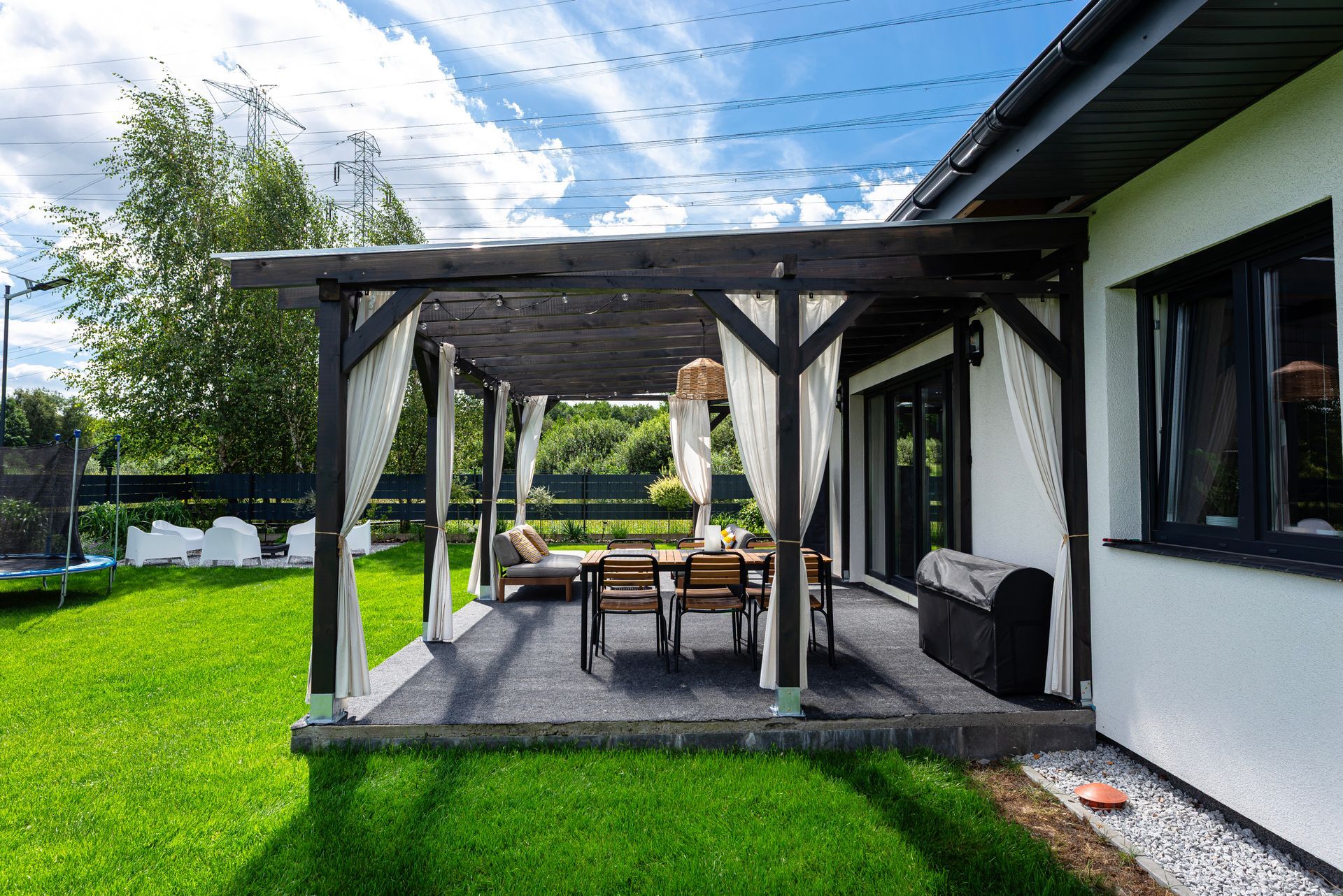 Covered backyard patio with wooden pergola, outdoor dining table, grill, and green lawn beside a house