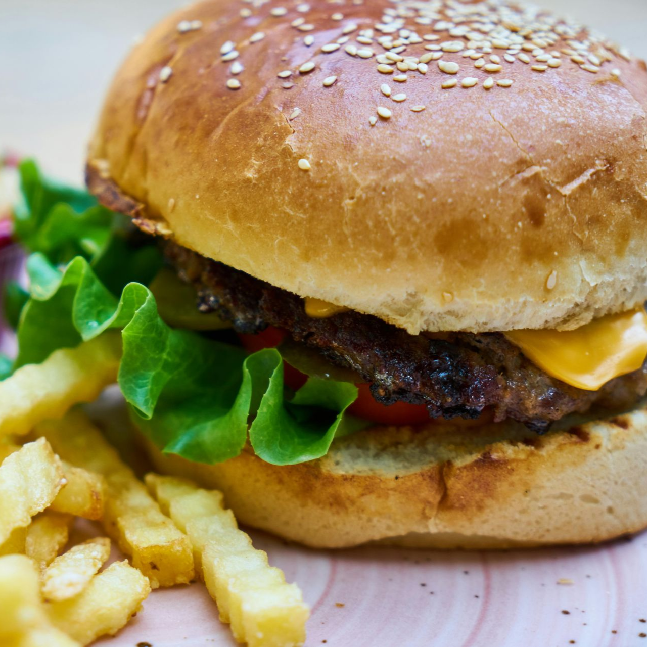 Hamburguesa con queso, lechuga, tomate y pepinillos en un pan con semillas de sésamo, servida con papas fritas.