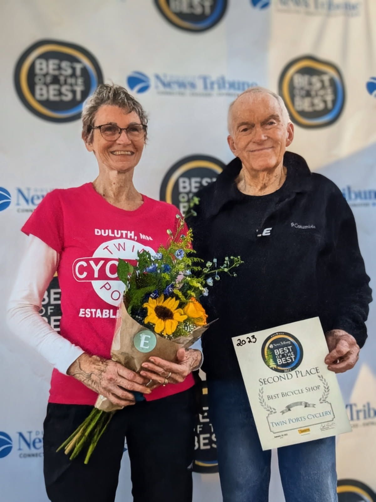 Woman in pink shirt and man in black jacket hold a bouquet and award, at a