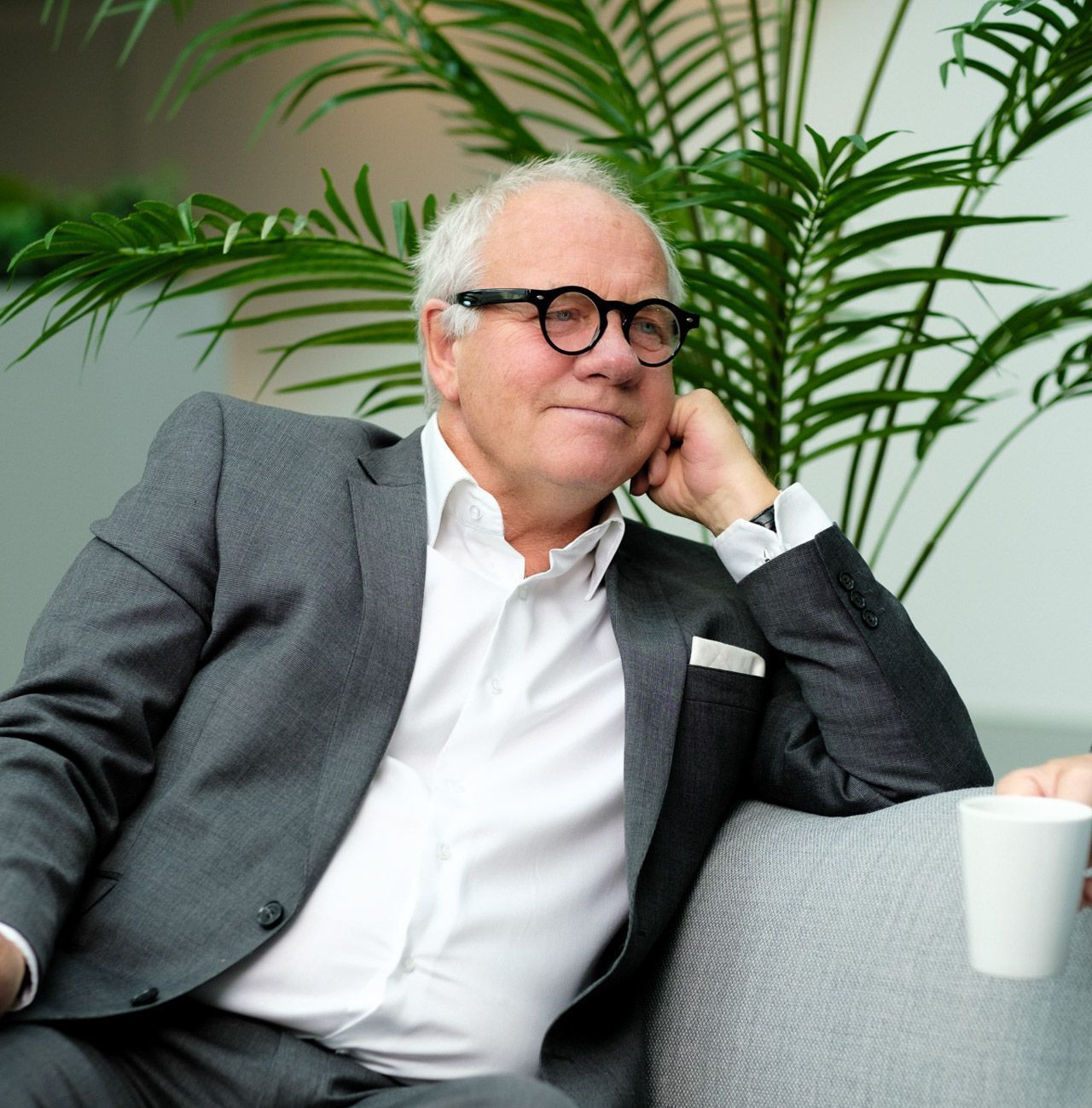 En mann i dress og briller sitter på en sofa.