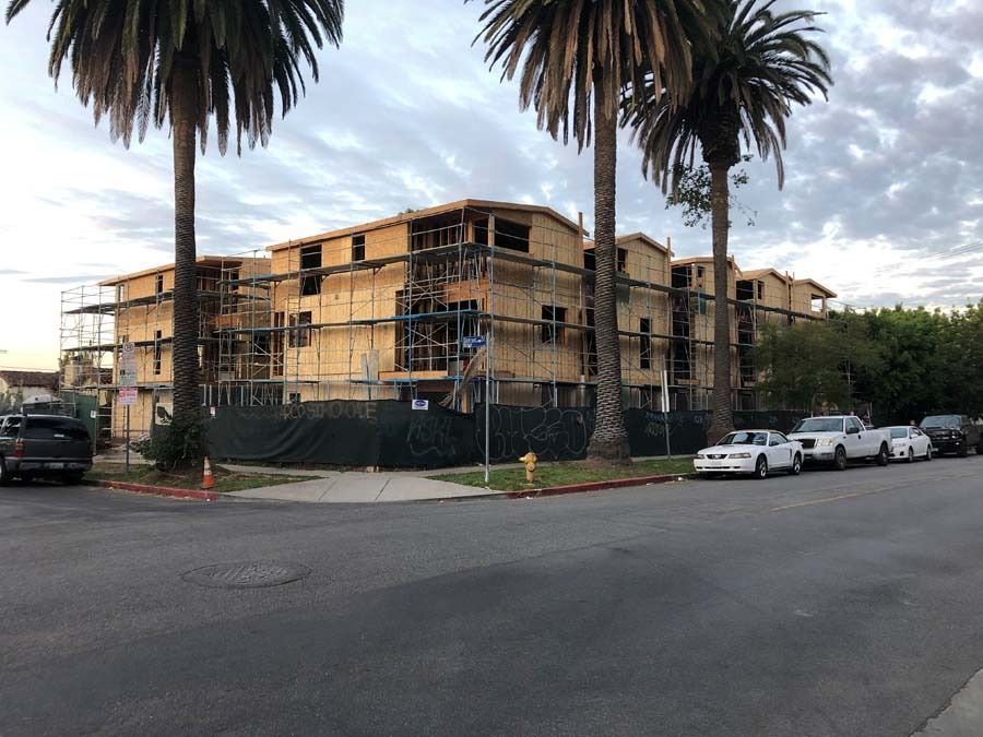 Construction site of a multi-story building with wooden frames. Palm trees and parked cars in front.