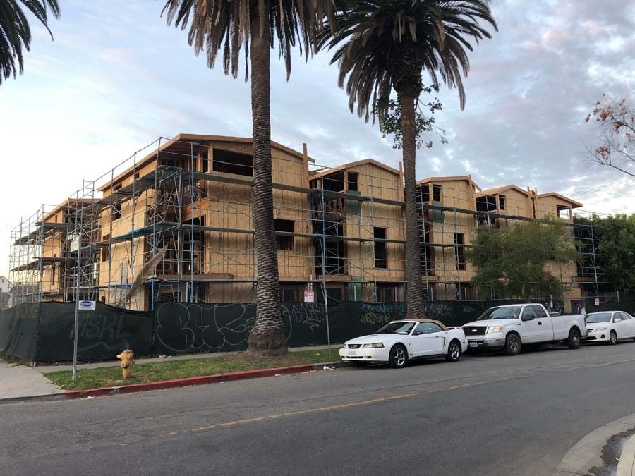 Construction site with a multi-story wooden building, scaffolding, palm trees, cars, and a street.
