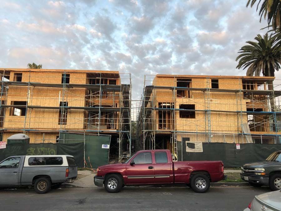 Two apartment buildings under construction, next to each other. Red and gray trucks parked in front, cloudy sky.