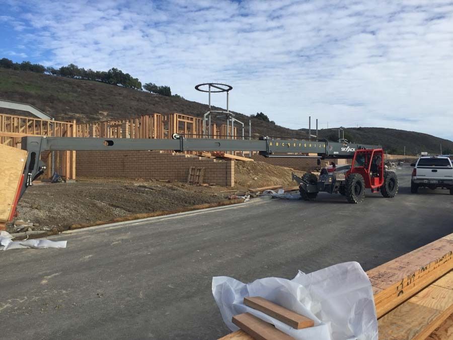 Construction site with a red forklift holding a long metal beam. Hills and unfinished houses are in the background.