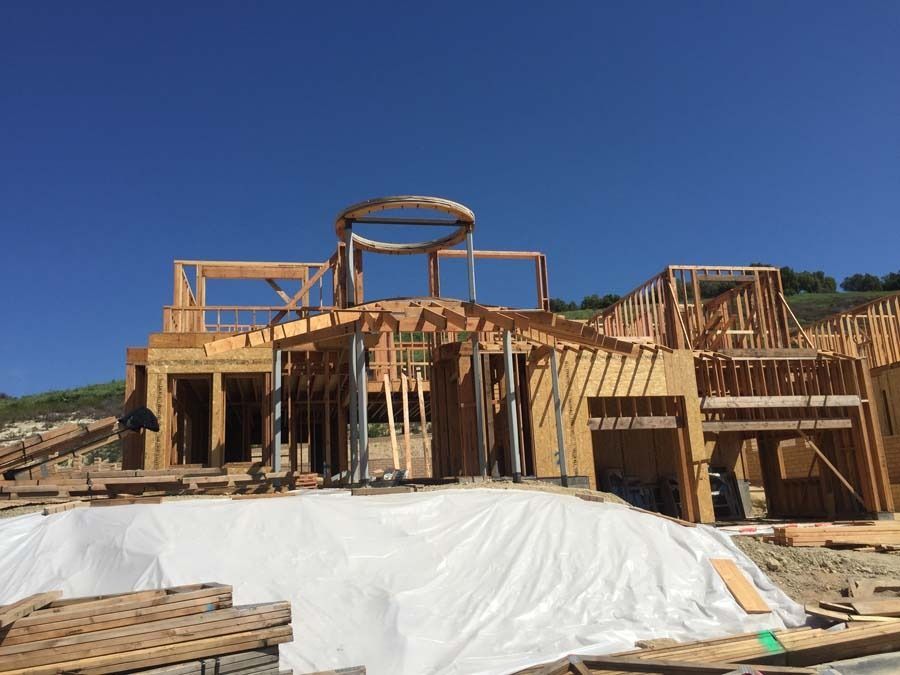 A house under construction with wooden framing, set against a blue sky. White tarp covers the ground in front.