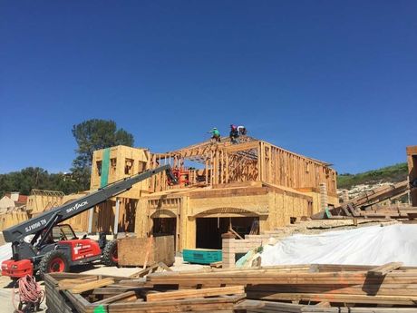 Construction workers building a wood-framed house under a clear blue sky; forklift in foreground.