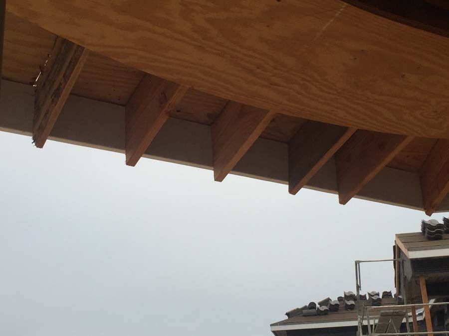 Wooden roof overhang with angled supports against a cloudy sky and a glimpse of a roof under construction.