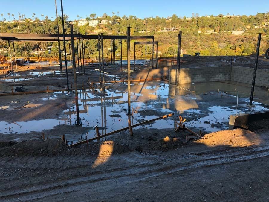 Construction site with standing water; metal beams and rebar frame a building foundation on a hillside.