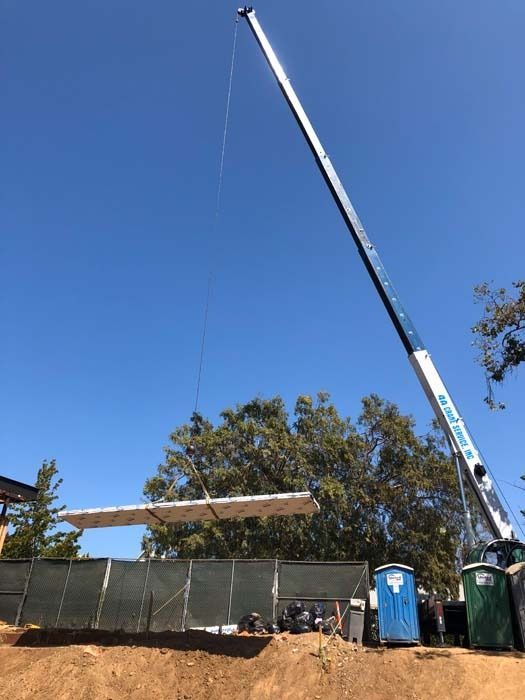 Crane lifting a long, rectangular beam above a construction site; blue sky and trees in the background.